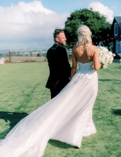 A bride and groom walk across a grassy field. The bride wears a white gown and holds a bouquet. The groom is in a black suit. A building and tree are in the background.