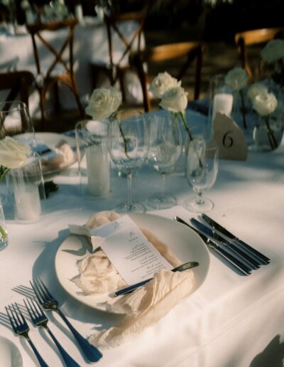 A table setting for a formal event with white plates, silver cutlery, menus, and glassware. White flowers and candles are arranged along the center of the table.