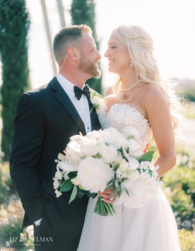 Bride and groom smiling at each other outdoors, bride holding a bouquet of white flowers.