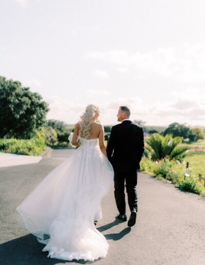 A bride in a white gown and a groom in a black suit walk hand in hand down a sunny outdoor path, surrounded by greenery.