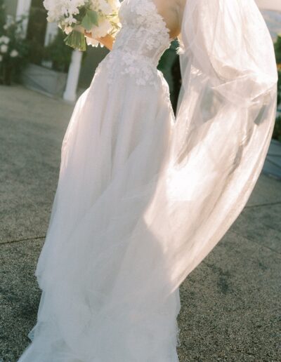 Bride in a white dress holding a bouquet, with a long flowing veil, standing outdoors on a sunny day.