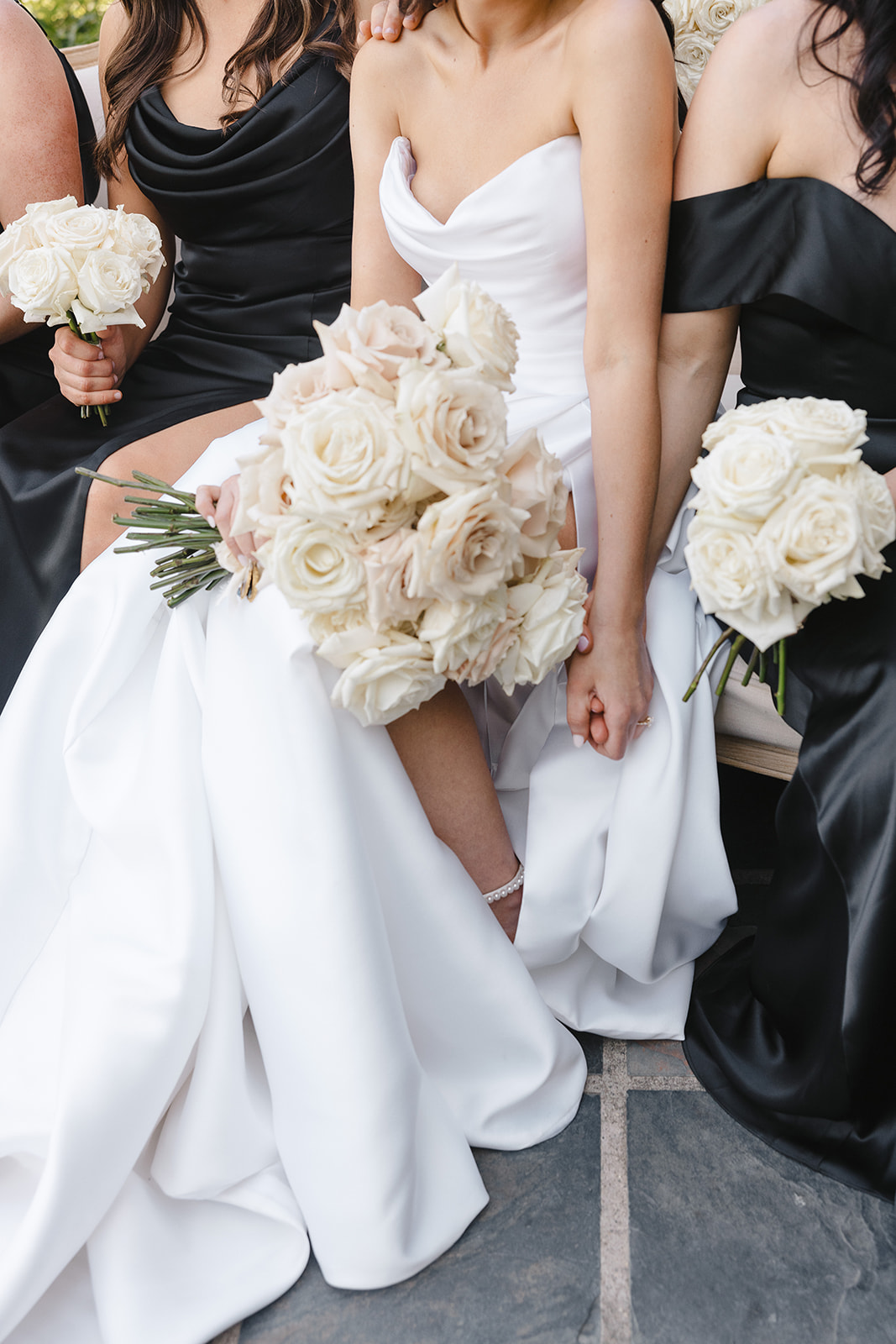 Bridesmaids in black dresses and a bride in a white gown hold bouquets of white roses while seated.