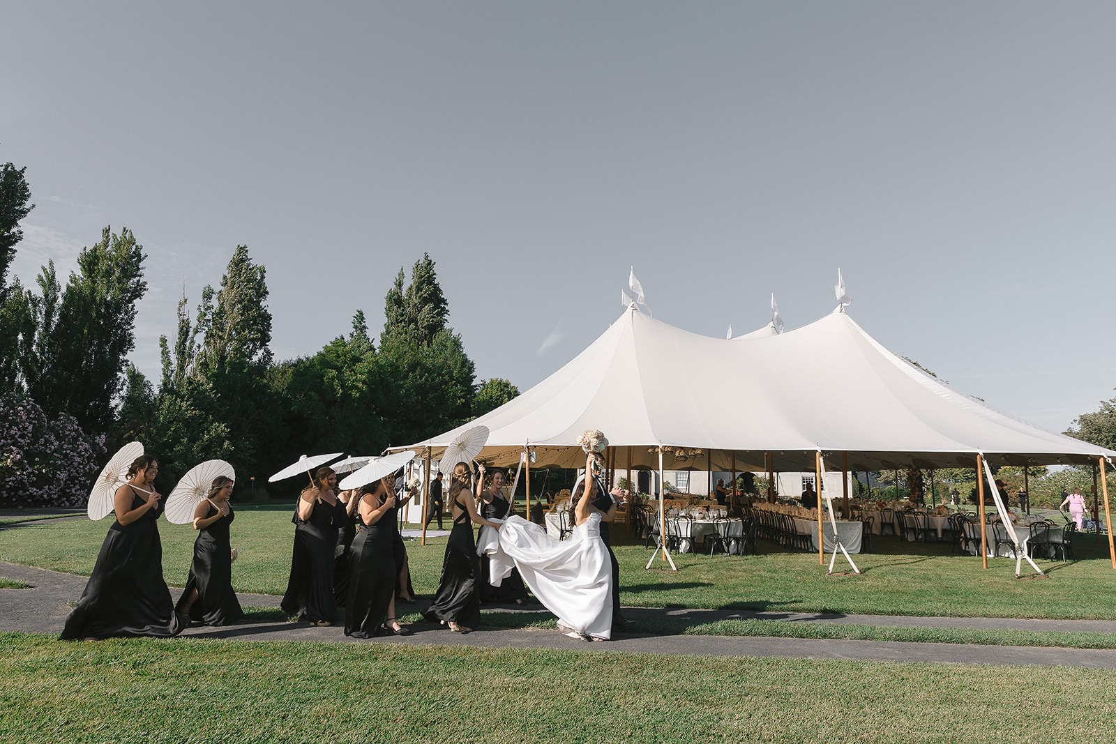 A bride in a white gown walks ahead of bridesmaids in black dresses holding parasols, near a large white tent on a grassy lawn, orchestrated flawlessly by a luxury Sacramento wedding planner.