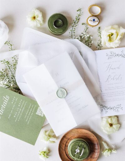 Elegant wedding invitation set with green wax seal, floral elements, and rings displayed on a white surface.