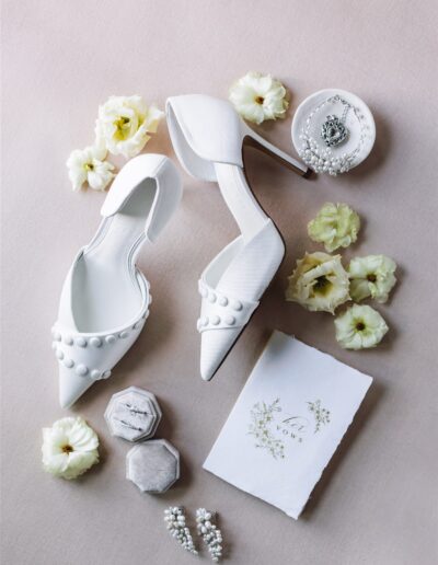 White bridal shoes with pearl details, surrounded by creamy flowers, a vows book, jewelry, and two small boxes on a soft background.