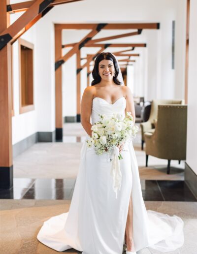 A bride in a white strapless gown holds a bouquet of white flowers while standing in a hallway.