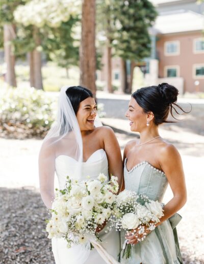 Two women stand outdoors, smiling at each other. One wears a white wedding dress and veil, holding a bouquet of white flowers. The other wears a light green dress, also holding a floral bouquet.