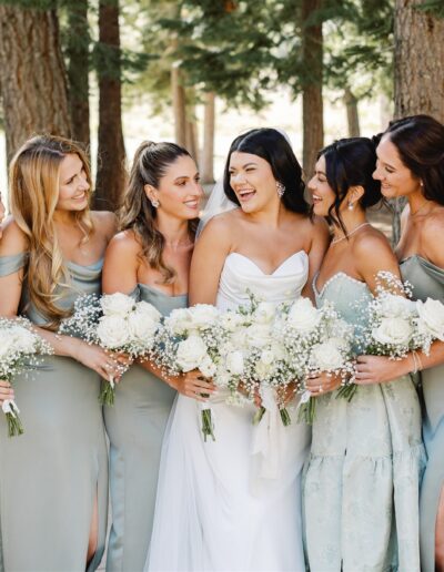 A bride in a white gown stands with six bridesmaids in matching sage dresses, holding bouquets of white flowers, surrounded by trees.