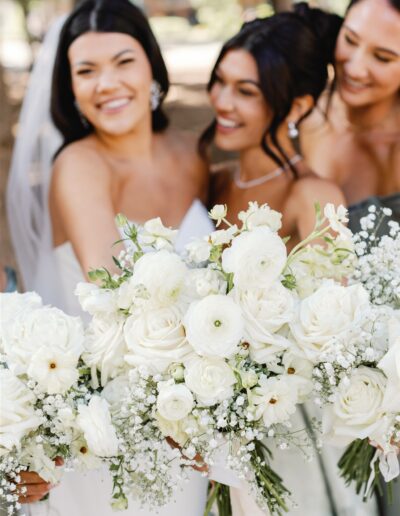 Three women in dresses hold white floral bouquets, smiling in an outdoor setting.