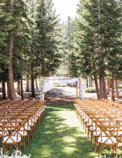 Outdoor wedding setup with wooden chairs lining an aisle leading to a floral arch among tall trees.