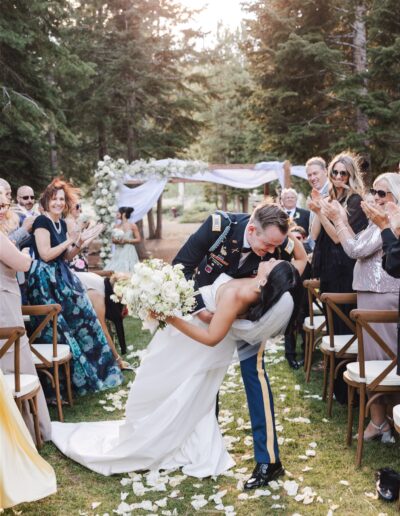 A bride and groom share a kiss at an outdoor wedding ceremony surrounded by guests, with petals scattered on the ground and an archway in the background.