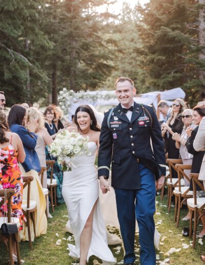 Bride and groom walking down the aisle outdoors, smiling, surrounded by guests and trees.