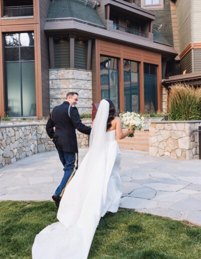 A couple walks hand in hand outside a building; the bride wears a white gown and veil, holding a bouquet, and the groom is in a military uniform.