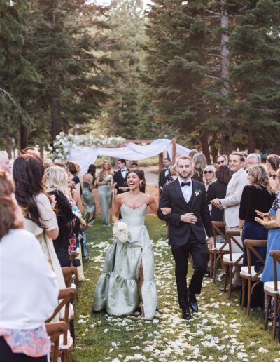 A bride and groom walk down an outdoor aisle lined with guests. The bride wears a green dress, and the groom is in a black tuxedo. They seem joyful. Trees and floral decorations surround the scene.