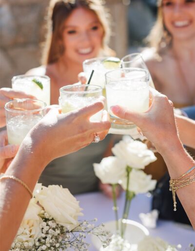 People clinking glasses in a toast, with white flowers on a table and a stone wall in the background.