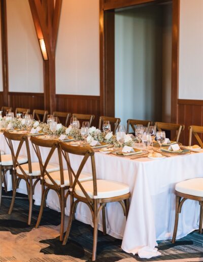 A long dining table is set with white cloth, floral centerpieces, and glassware, surrounded by wooden chairs in a warmly lit room.