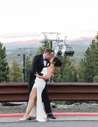 A couple dressed in formal attire shares a dance on a roadside with cable cars and pine trees in the background.