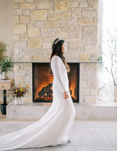 A person in a long white dress walks across a room with a stone fireplace in the background.