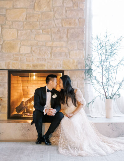 A couple in formal attire sits by a stone fireplace, leaning in close, with a vase of greenery nearby.