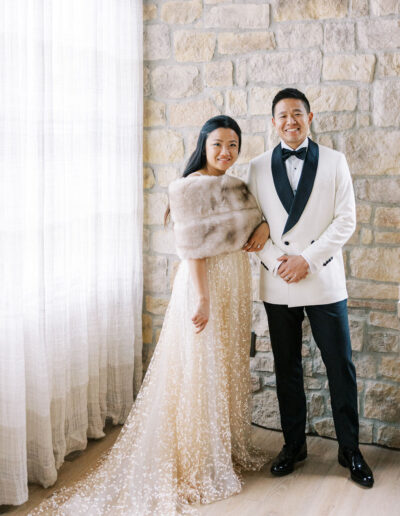 Couple posing indoors; woman in a sparkly gown with a fur stole and man in a white tuxedo jacket and black pants, standing against a stone wall next to a sheer curtained window.