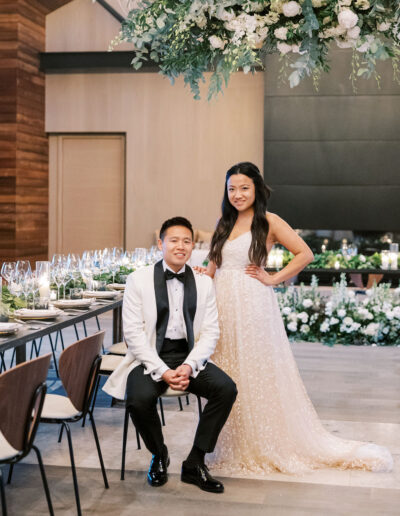 A couple in formal attire poses in an elegantly decorated room with a table set for an event.