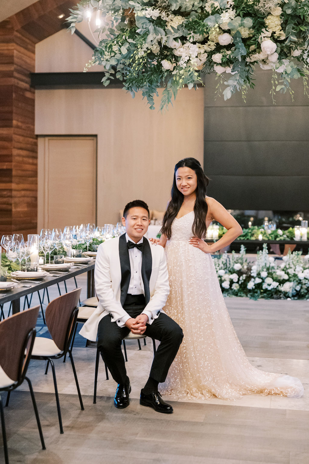 A couple in formal attire pose in a decorated room with a dining setup, featuring floral arrangements and elegant table settings.