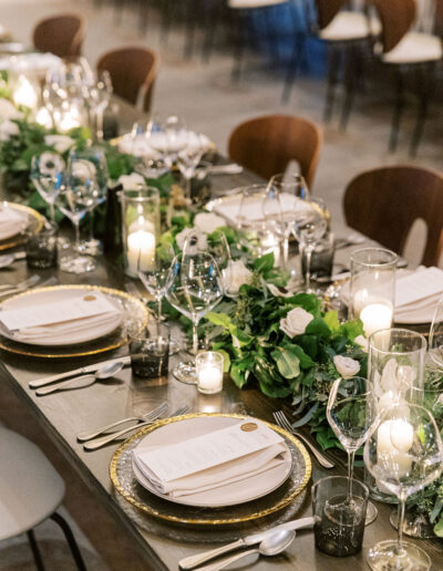 Elegant dining table set with gold-rimmed plates, cutlery, wine glasses, candles, and greenery centerpieces.