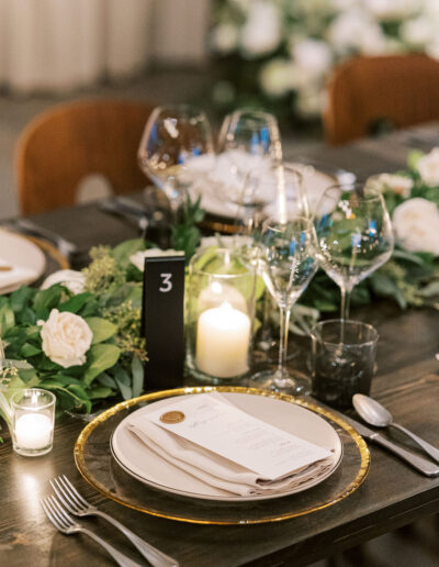 Elegant table setting with a plate, napkin, menu, and cutlery on a dark wooden table. Wine glasses, candles, and greenery adorn the table. A black table number "3" is visible.