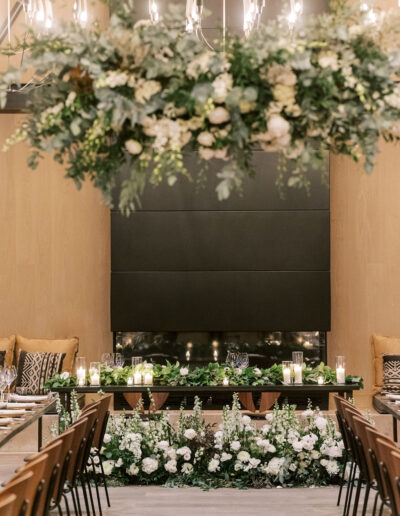 Elegant indoor wedding setting with a floral arch above a table, surrounded by candles and white flowers, against a black wall backdrop. Seats arranged facing the table.