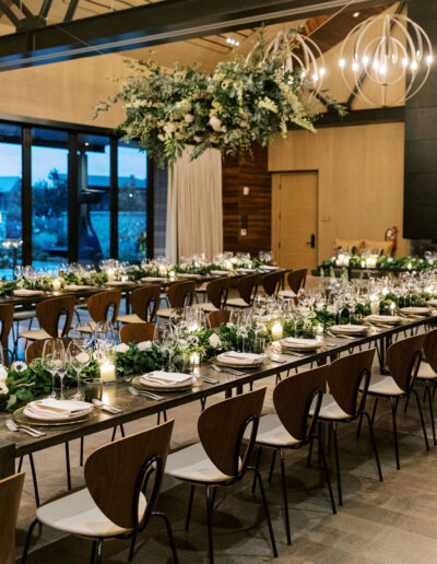 Elegantly set dining tables with glassware, plates, and greenery centerpieces under modern chandeliers in a spacious room.