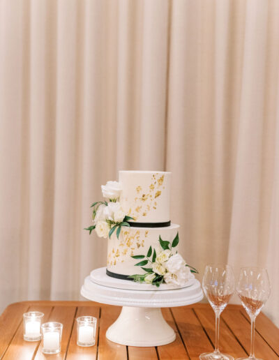 Two-tier white cake with gold accents and white flowers on a stand. Three lit candles and two empty wine glasses are on a wooden table, set against a beige curtain backdrop.