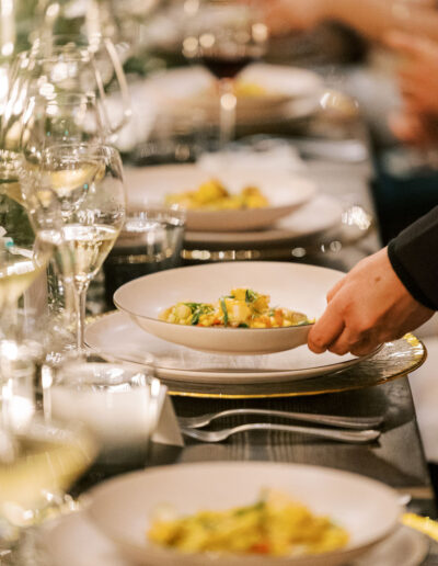 A hand places a dish of pasta on a elegantly set dining table, surrounded by plates and wine glasses.