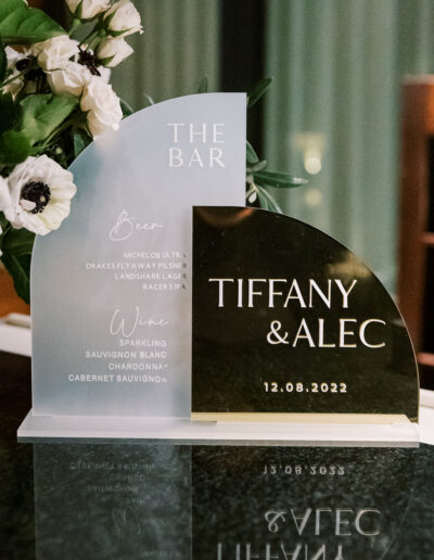 A wedding bar sign with selected beer and wine offerings beside white flowers. The names "Tiffany & Alec" and date "12.08.2022" are displayed.