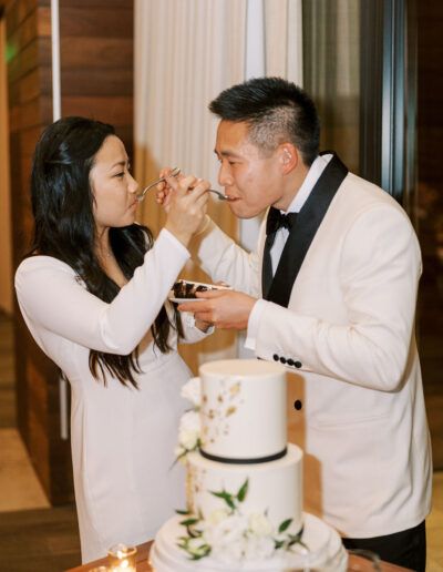 A couple in formal attire shares a piece of cake. A two-tier cake and lit candles are in the foreground. They are indoors.