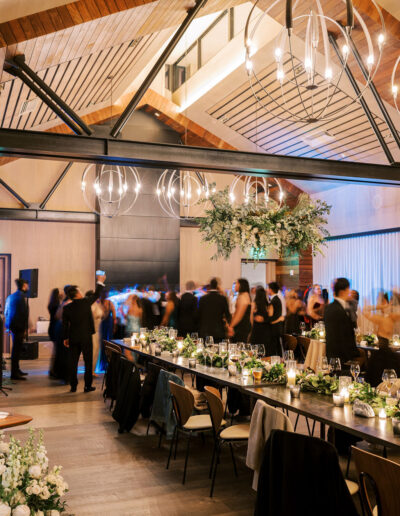 A large dining room with long tables set for a formal event. People are mingling under modern chandeliers and a high wooden ceiling.