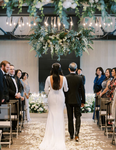 A bride and an older man walk down an aisle covered with white petals, surrounded by seated guests, under a large floral chandelier.