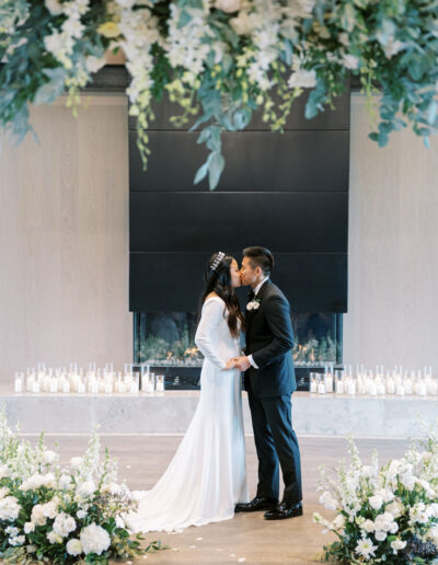 A bride and groom kiss at their wedding ceremony, framed by floral arrangements and white candles.