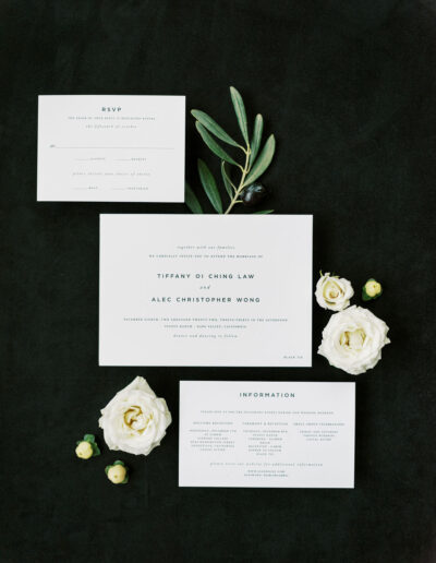 Wedding invitation set with RSVP and information cards, adorned with white flowers and green leaves on a dark background.