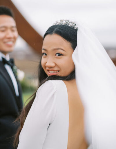 Bride in a white dress smiles at the camera with a groom in a black suit in the background. They are outdoors with a blurred, natural setting.