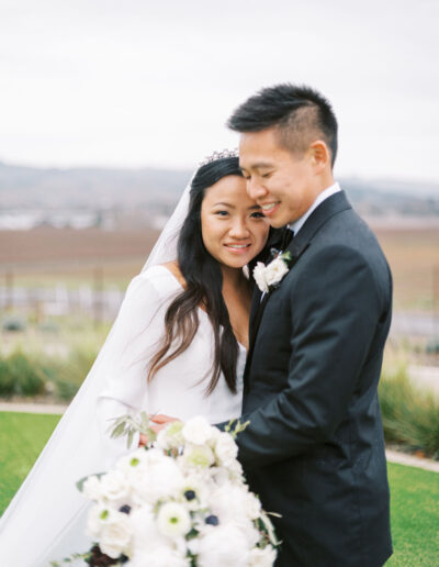 A couple in wedding attire stands outdoors. The bride holds a bouquet of white flowers. The background features grass and distant hills under a cloudy sky.