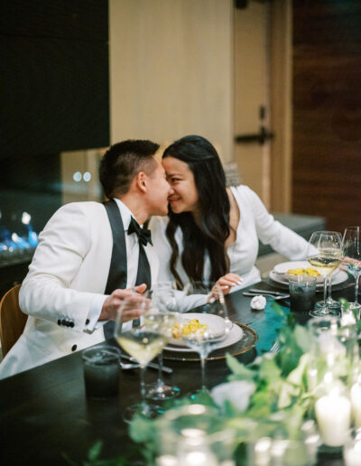 A couple in formal attire leans towards each other, smiling, while seated at a dinner table with plates, wine glasses, and candles.