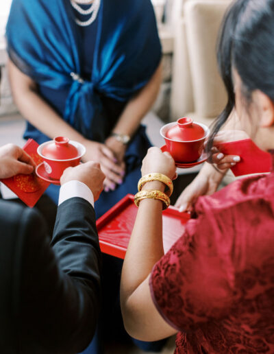 A woman and man hold red tea cups and red envelopes, participating in a traditional tea ceremony. A seated woman in a blue shawl watches.