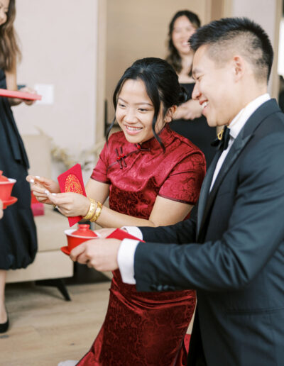 A couple in traditional attire is kneeling, holding red envelopes and teacups, surrounded by others during a tea ceremony.