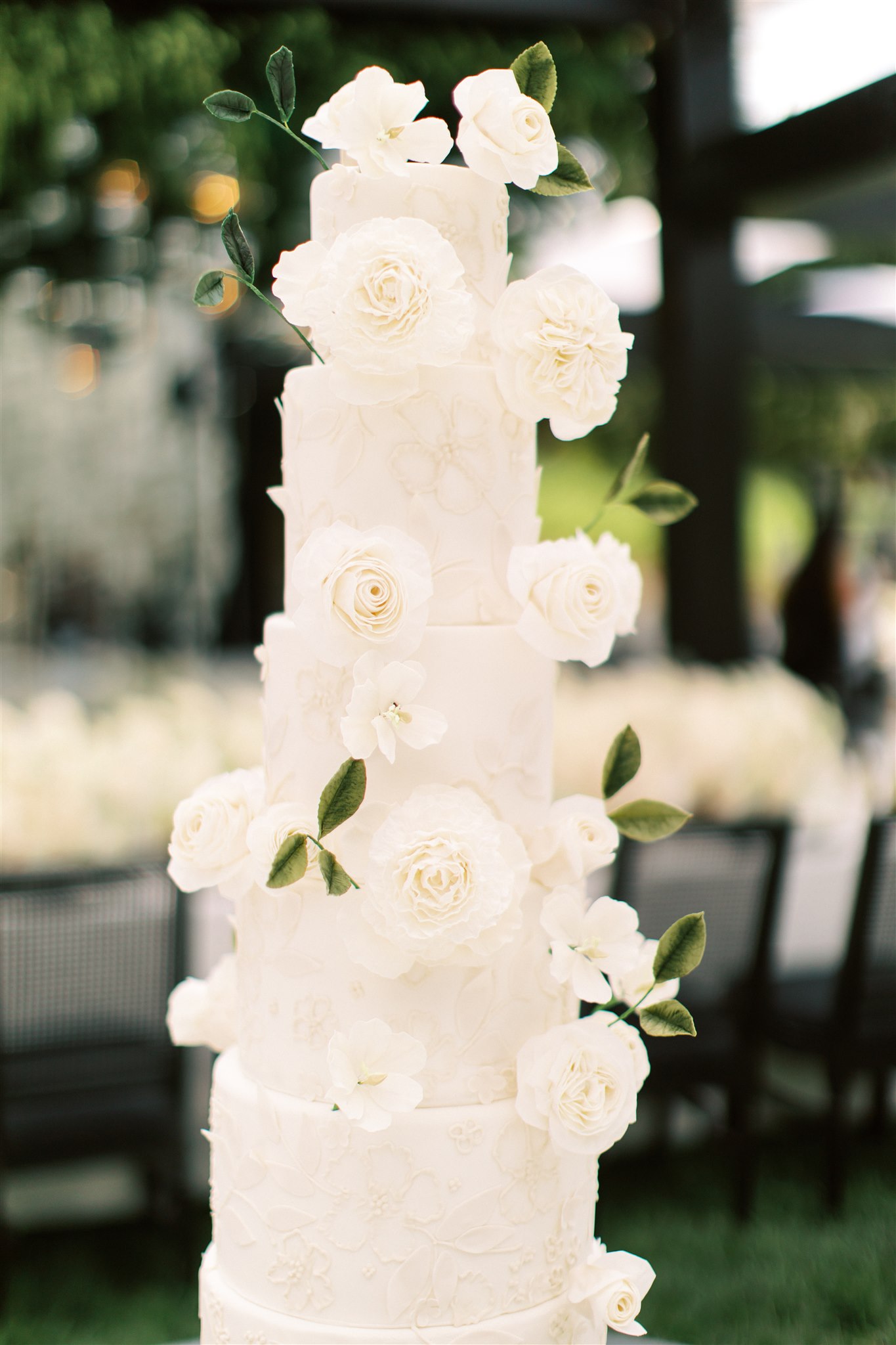 A tall white wedding cake adorned with large white roses and green leaves, set against an outdoor backdrop.