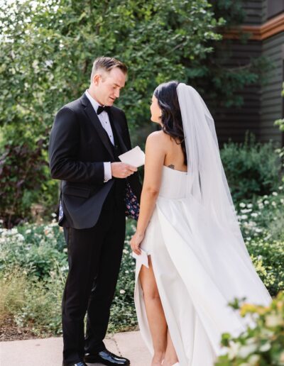 A bride and groom stand facing each other outdoors. The groom is holding a folded piece of paper, and the bride is wearing a white dress with a veil. They are surrounded by greenery.