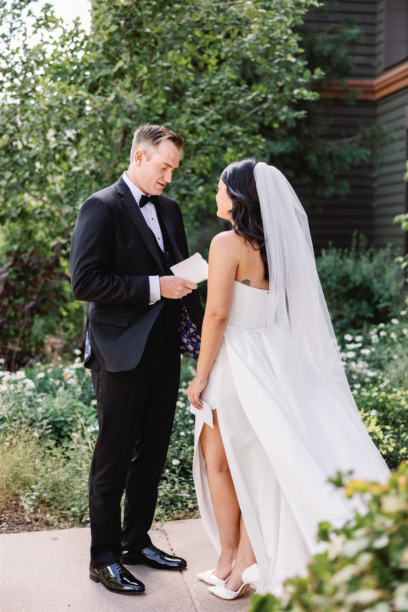 A bride and groom stand facing each other outdoors. The groom is holding a folded piece of paper, and the bride is wearing a white dress with a veil. They are surrounded by greenery.