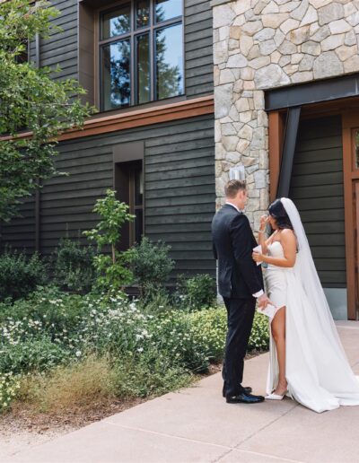 A couple in wedding attire stands holding hands in front of a building with stone and wood siding, surrounded by greenery and flowers.