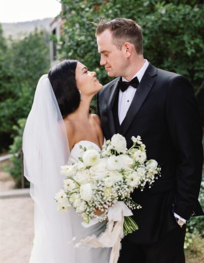 A bride and groom share a loving gaze outdoors. The bride holds a bouquet of white flowers and wears a veil. The groom is in a black suit and bowtie. Greenery and buildings are in the background.