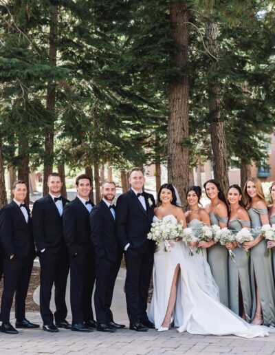 A wedding party poses in a wooded outdoor setting. The group includes bridesmaids in matching light green dresses and groomsmen in black suits.