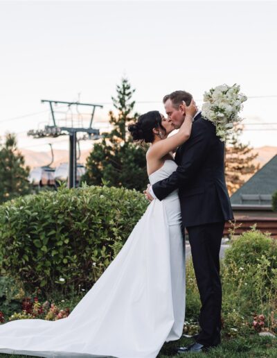 Bride and groom kissing in a garden setting with mountains and a lodge in the background.